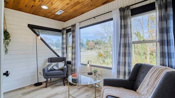 Une pièce avec un plafond en bois, de grandes fenêtres avec des rideaux, un canapé, un fauteuil et une petite table.
