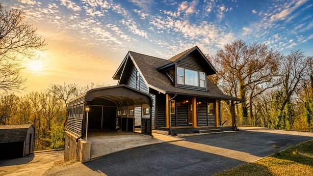 A two-story log cabin with a covered garage, a large driveway, and a scenic view of trees and sky. - The Black Log Cabin - Hot Tub & Game Room (Fredericksburg)