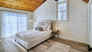 Une chambre avec un plafond en bois, un lit avec des oreillers, une table de chevet, une lampe sur pied et une fenêtre.