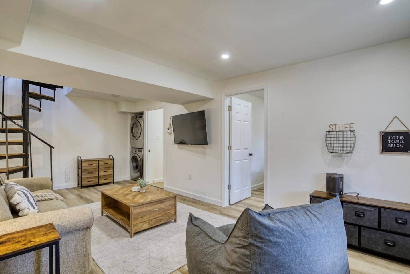 A modern living room with a sofa, coffee table, and a TV. There's a staircase and a laundry area visible.