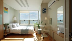 Une chambre moderne avec un grand lit, un bureau et une chaise, une plante en pot, et une vue sur la ville grâce à de grandes fenêtres.