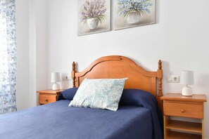 Ein Schlafzimmer mit einem Holz-Kopfteil, einer blauen Bettdecke, einem floralen Kissen, zwei Nachttischen mit Lampen und zwei an der Wand hängenden floralen Bildern.
