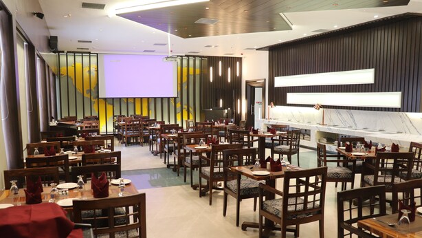 A spacious dining area with multiple tables and chairs, a stage with a projection screen, and a modern bar area. - Hotel Windsor (Patna)