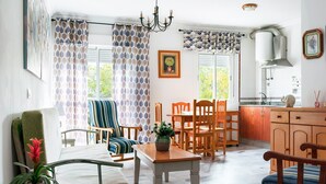 A living room with a dining table, chairs, a sofa, a potted plant, and a kitchen area with cabinets and a sink.