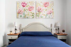 A bedroom with a white headboard, a blue bedspread, two bedside tables with lamps, and two floral paintings on the wall.