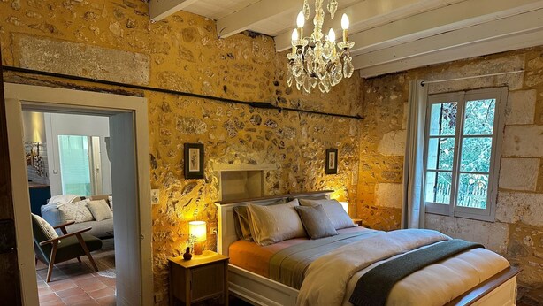 Un dormitorio con pared de piedra, un candelabro, una cama con almohadas, una mesita de noche con una lámpara y una ventana con vistas a árboles.