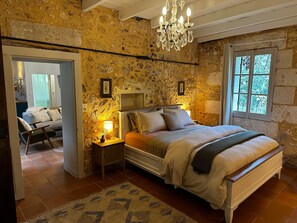 Une chambre avec un mur en pierre, un lustre, un lit avec des oreillers, une table de chevet avec une lampe et une fenêtre donnant sur des arbres.
