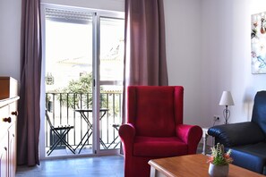 Een kamer met een rode stoel, een blauwe bank, een houten tafel met een plant en een balkon met een tafel en stoelen.