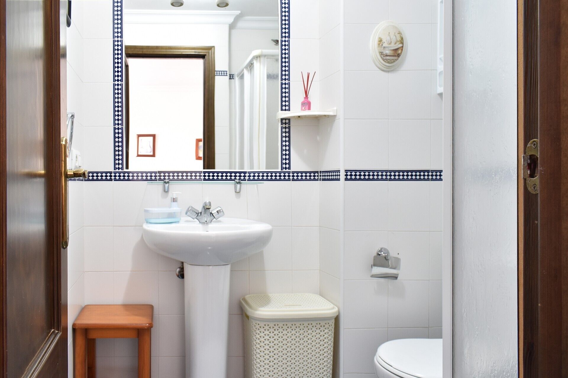 Un baño con un lavabo blanco, un espejo, un inodoro, un taburete de madera pequeño y un cubo de basura.