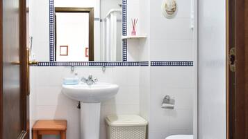 Un baño con un lavabo blanco, un espejo, un inodoro, un taburete de madera pequeño y un cubo de basura.