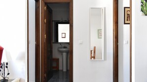 Passadís que condueix a un bany amb un lavabo i mirall, un tamboret petit i un quadre al mur.