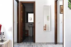Un pasillo que conduce a un baño con lavamanos y espejo, un taburete pequeño y un cuadro en la pared.