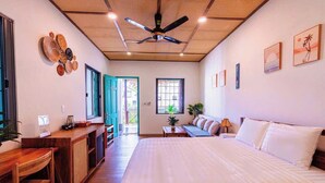 Une chambre spacieuse avec un grand lit, un bureau en bois, un ventilateur de plafond et une vue dégagée sur l’extérieur grâce à une porte ouverte.