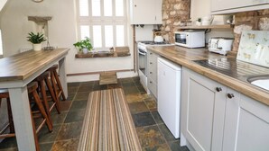 Una cocina con encimera de madera, armarios blancos, salpicadero de piedra, una ventana con persianas y un reloj en la pared.