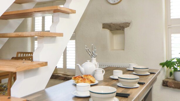 Un comedor con una escalera de madera, un reloj en la pared y una mesa servida con platos, tazones y una tetera.