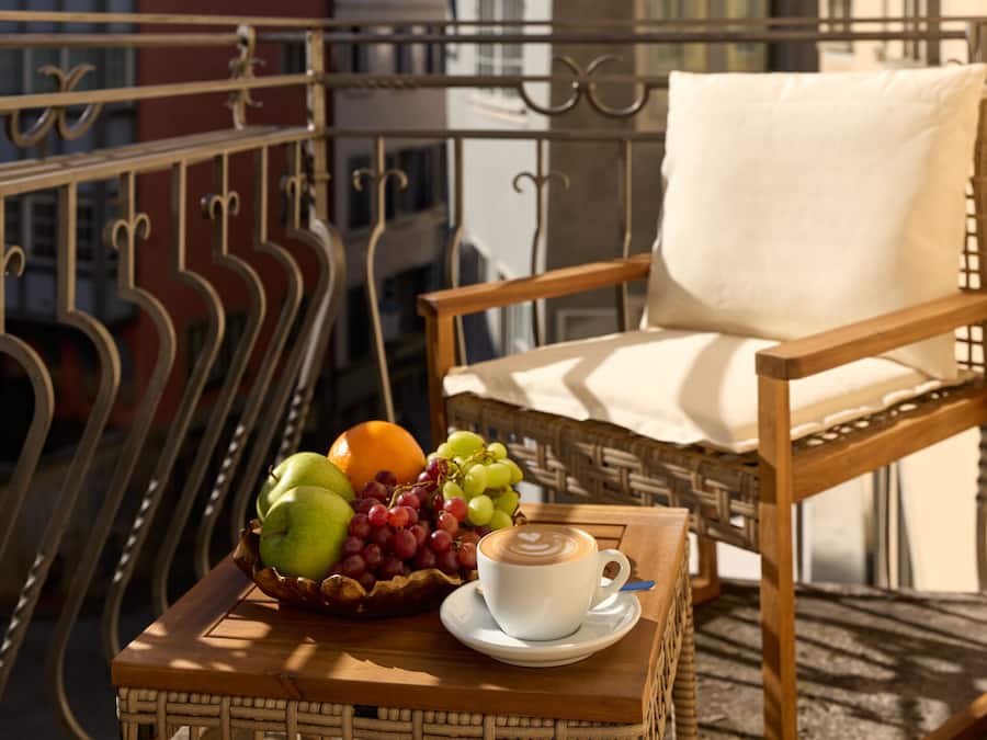 Uma cadeira de vime com almofada, uma mesinha com uma fruteira, uma xícara de café e um corrimão de varanda.