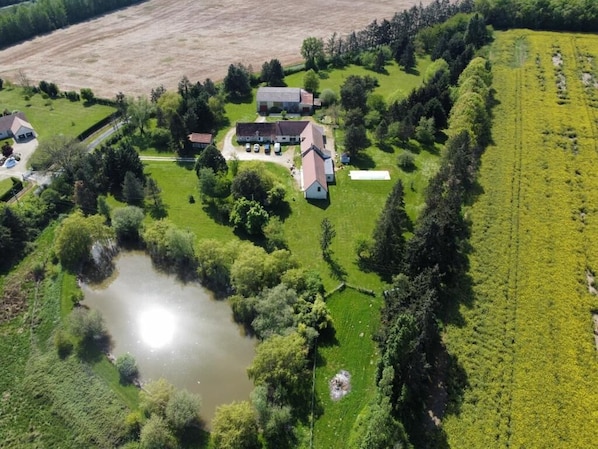 Oversigtsbillede af en landejendom med et stort hus, en sø og marker i det omkringliggende landskab.