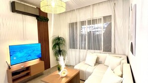 Ein modernes Wohnzimmer mit einem Flachbildfernseher, einem Holztisch, einem weißen Sofa und einem großen Fenster mit transparenten Vorhängen.