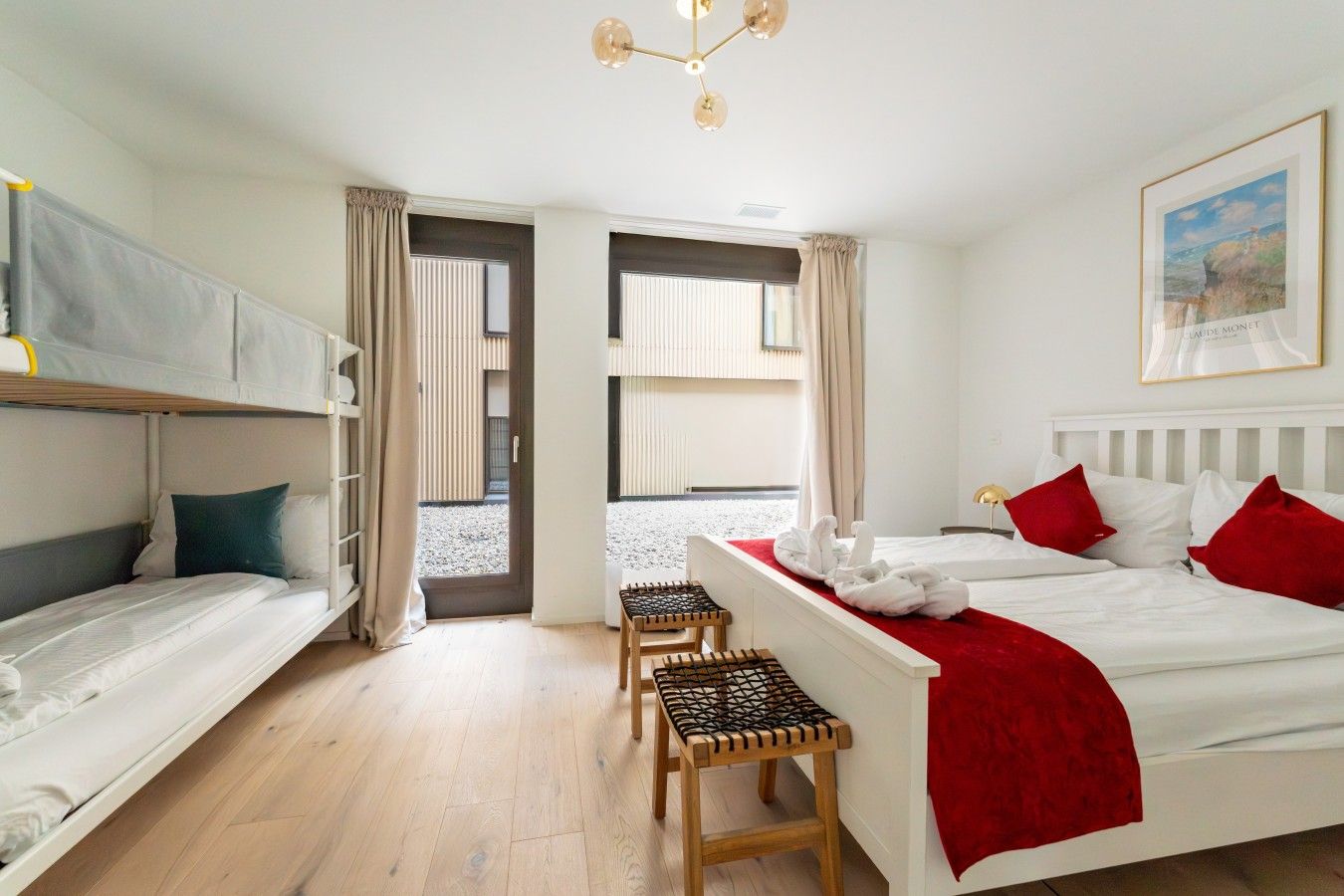 Une chambre d’hôtel avec un lit superposé, un lit simple, un banc en bois et une vue sur l’extérieur par une porte ouverte.