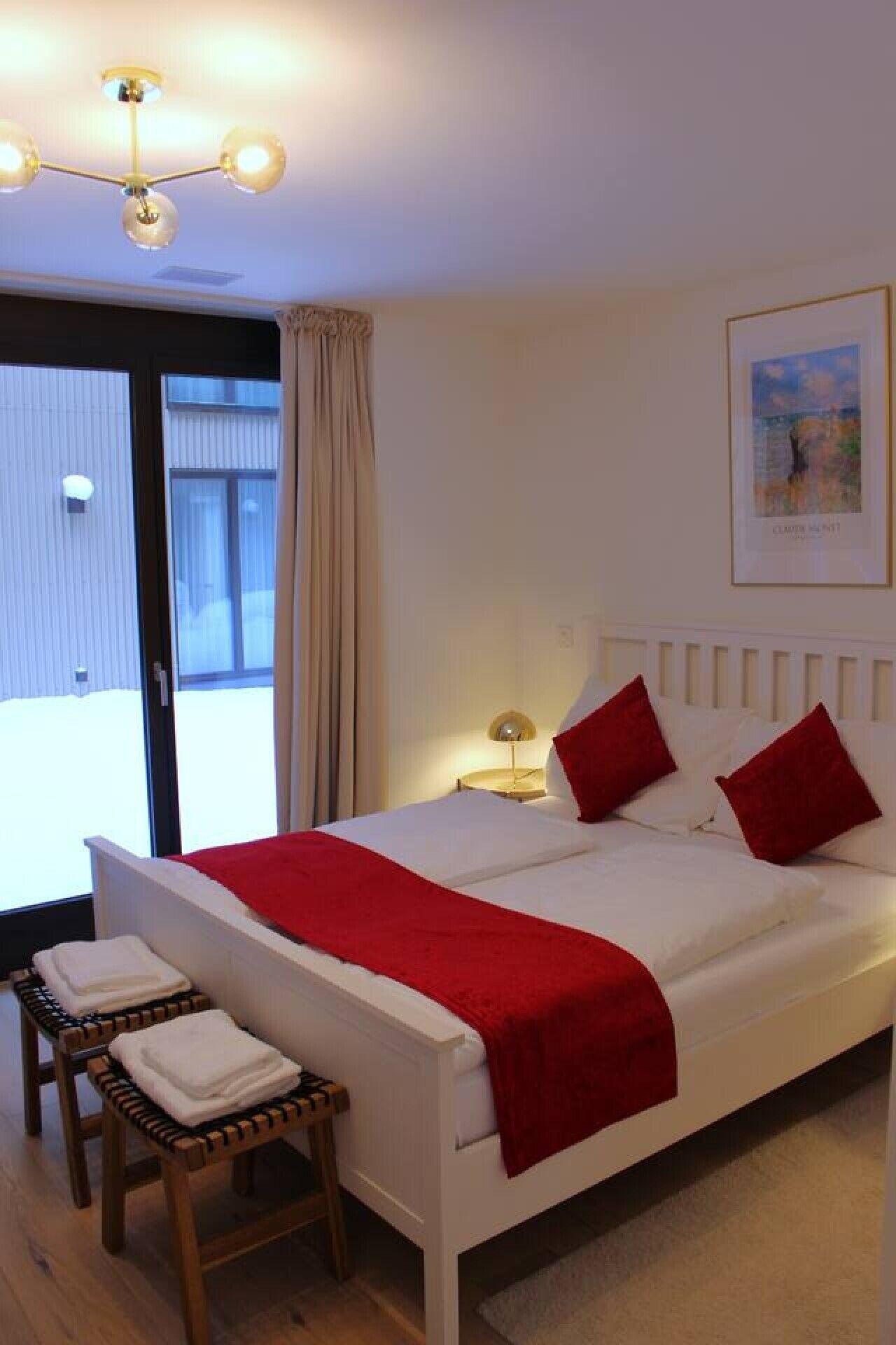 A bedroom with a bed, pillows, a bedside table, a lamp, a window with curtains, and a framed picture on the wall.