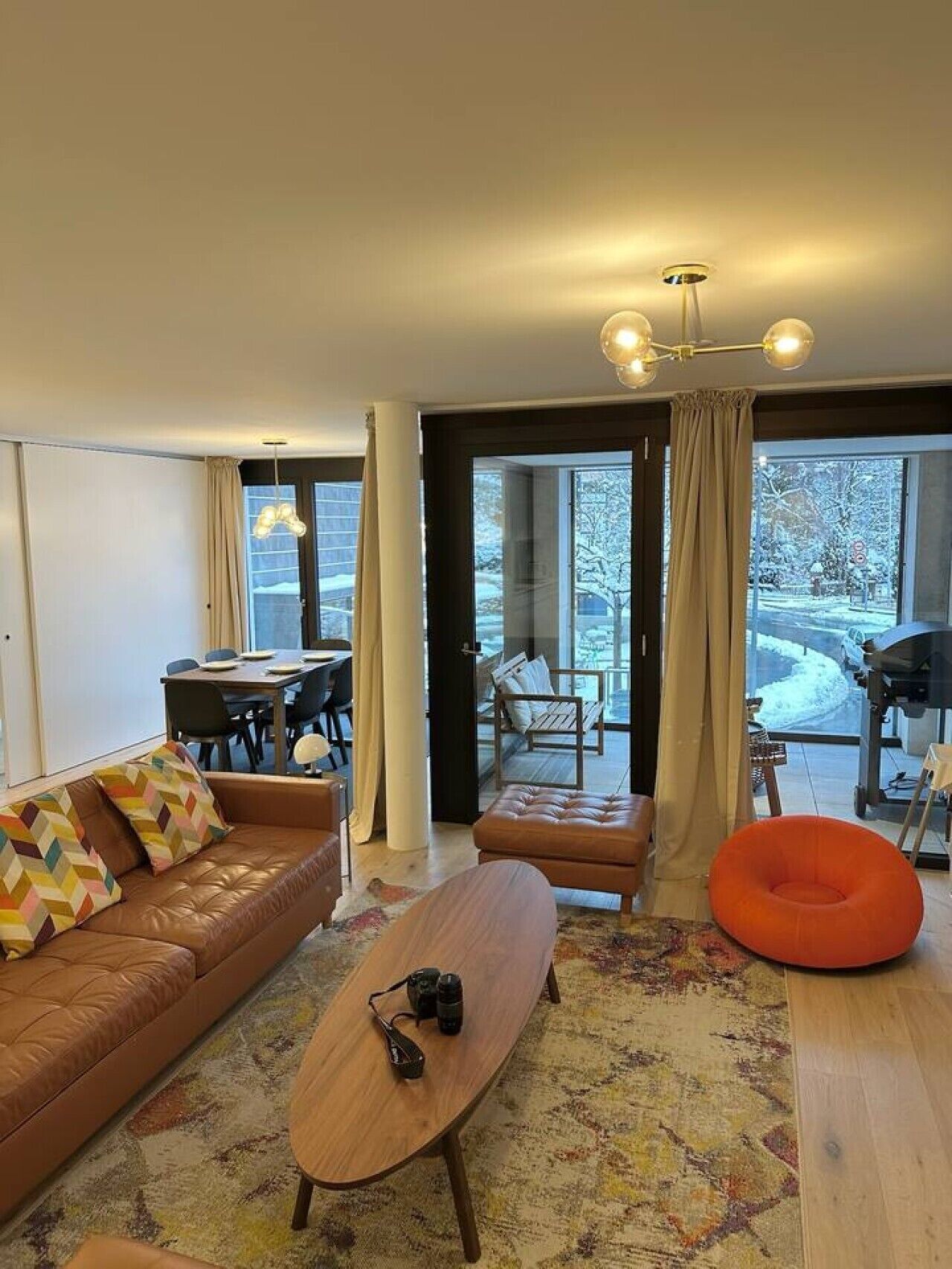Ein modernes Wohnzimmer mit einer Couch, einem Couchtisch und einem orangenen Sitzsack. Es gibt einen Essbereich mit Tisch und Stühlen sowie einen Blick ins Freie durch große Fenster.