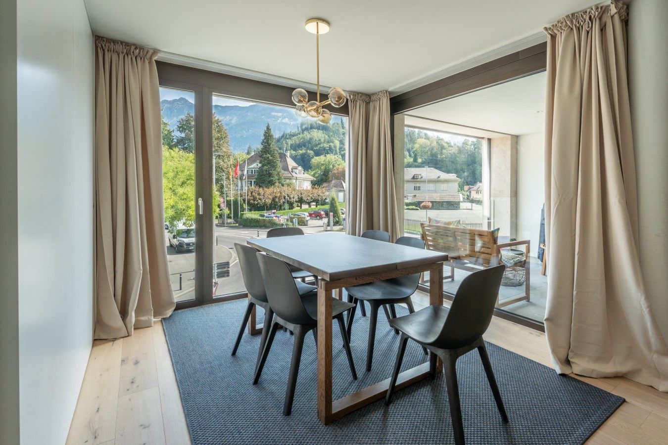 Un comedor con mesa y sillas, ventanas amplias con cortinas y vistas a un paisaje montañoso.