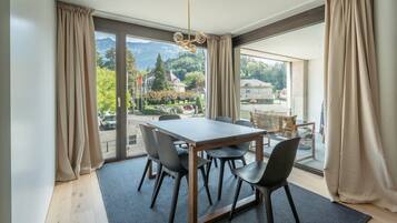 Un comedor con mesa y sillas, ventanas amplias con cortinas y vistas a un paisaje montañoso.