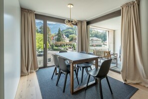 Ein Essbereich mit Tisch und Stühlen, große Fenster mit Vorhängen und Blick auf eine Berglandschaft.