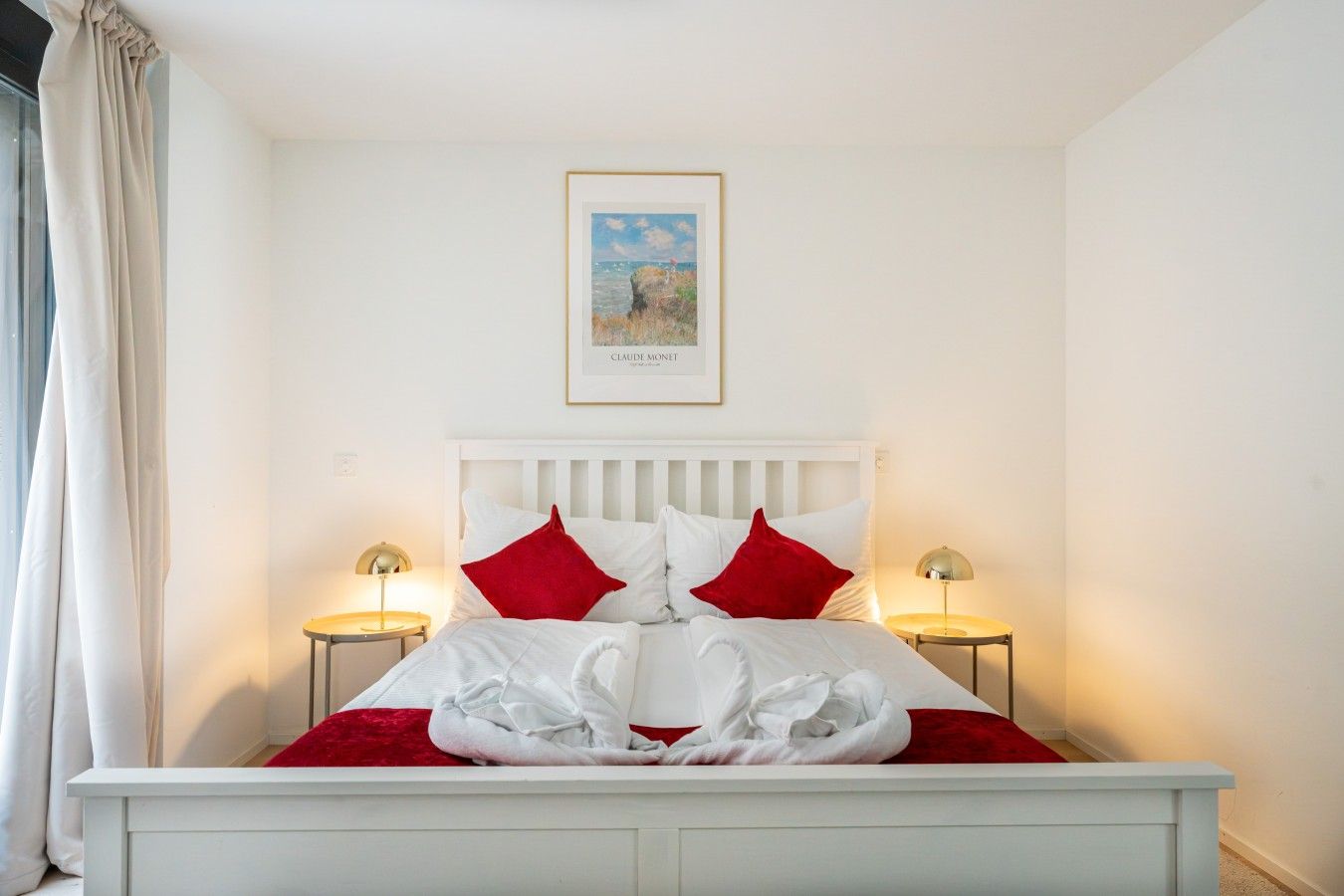 A neatly made bed with white bedding and red pillows, two bedside lamps, a framed artwork above the bed, and a white headboard.