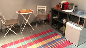 Eine kompakte Küchenzeile mit Mikrowelle, Toaster und kleinem Kühlschrank. Ein Klappsessel und ein kleiner Tisch mit Tablett sind vorhanden.