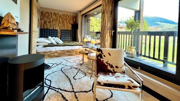 Een moderne slaapkamer met een groot raam, een bed met houten hoofdeinde, een bank, een stoel en een tafeltje met een lamp.