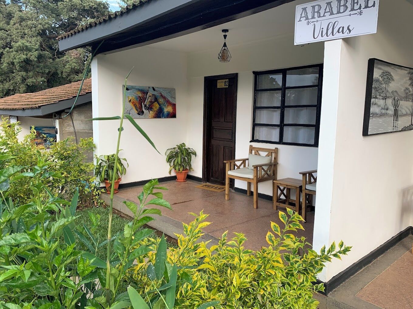 A villa with a sign reading "ARABEL Villas," a wooden chair, potted plants, and a painting on the wall.