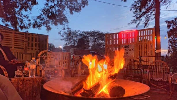 A campfire with logs and flames, surrounded by wooden chairs and a table with bottles. - Riverside Villas Nanyuki (Nanyuki)