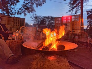 A campfire with logs and flames, surrounded by wooden chairs and a table with bottles. - Riverside Villas Nanyuki (Nanyuki)