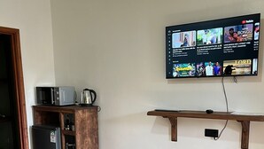 A hotel room with a wooden desk, a microwave, a refrigerator, and a TV mounted on the wall displaying music videos. - Riverside Villas Nanyuki (Nanyuki)