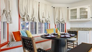 Un comedor con una mesa redonda, sillas y un banco. Hay cortinas atadas, un mueble con puertas de cristal y una ventana con persianas.
