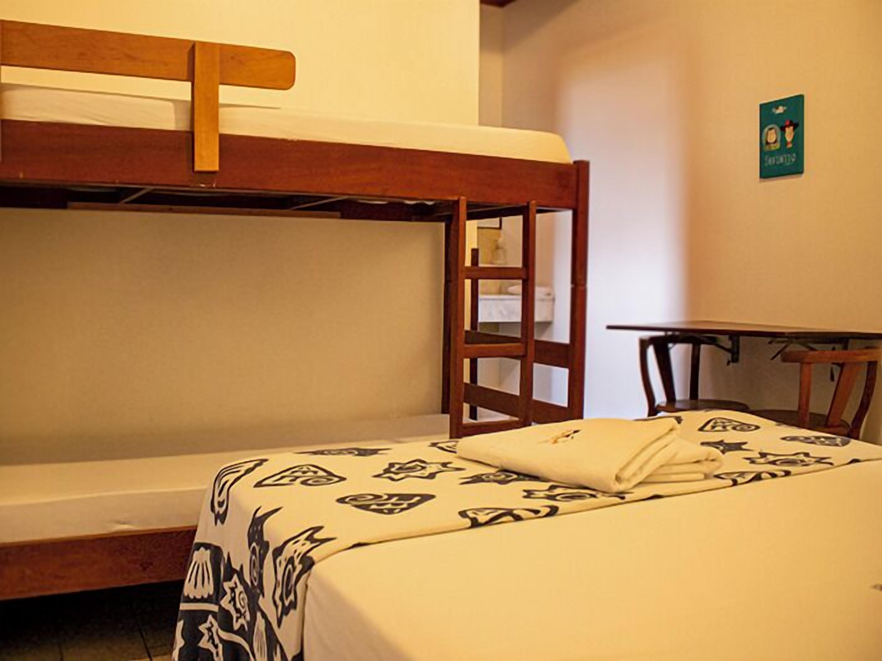Una cama individual con tablero de madera, una mesita pequeña y una silla en una habitación con paredes lisas.