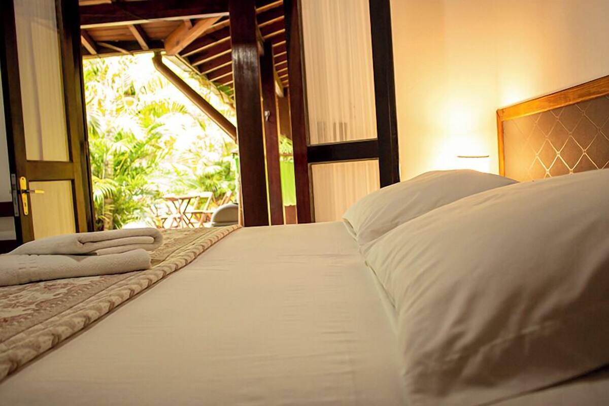 A neatly made bed with white linens and pillows, a wooden headboard, and a view of an outdoor area with greenery.