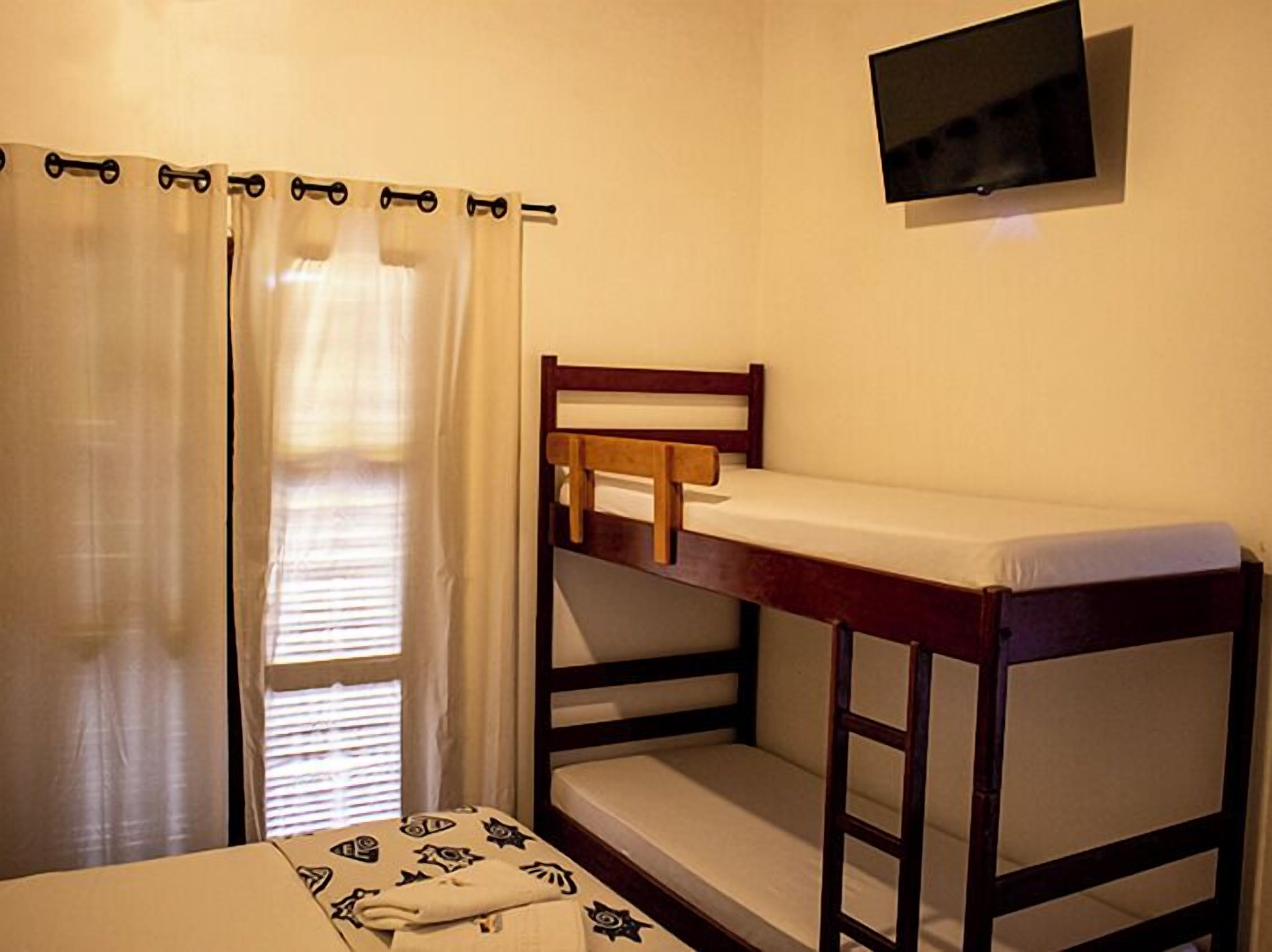 Quarto com beliches, televisão presa à parede e janela com cortinas.
