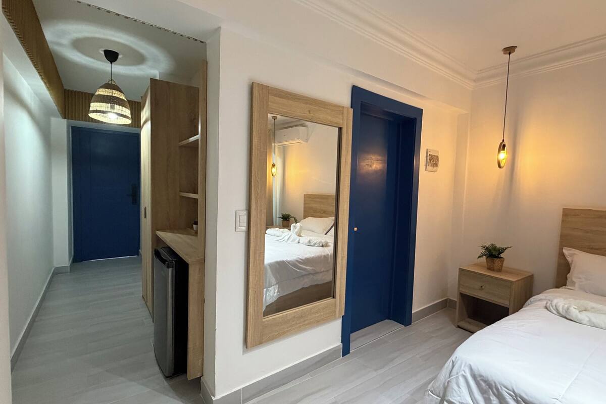 A hotel room with a bed, bedside table, and a mirror. There is a hallway leading to another room.