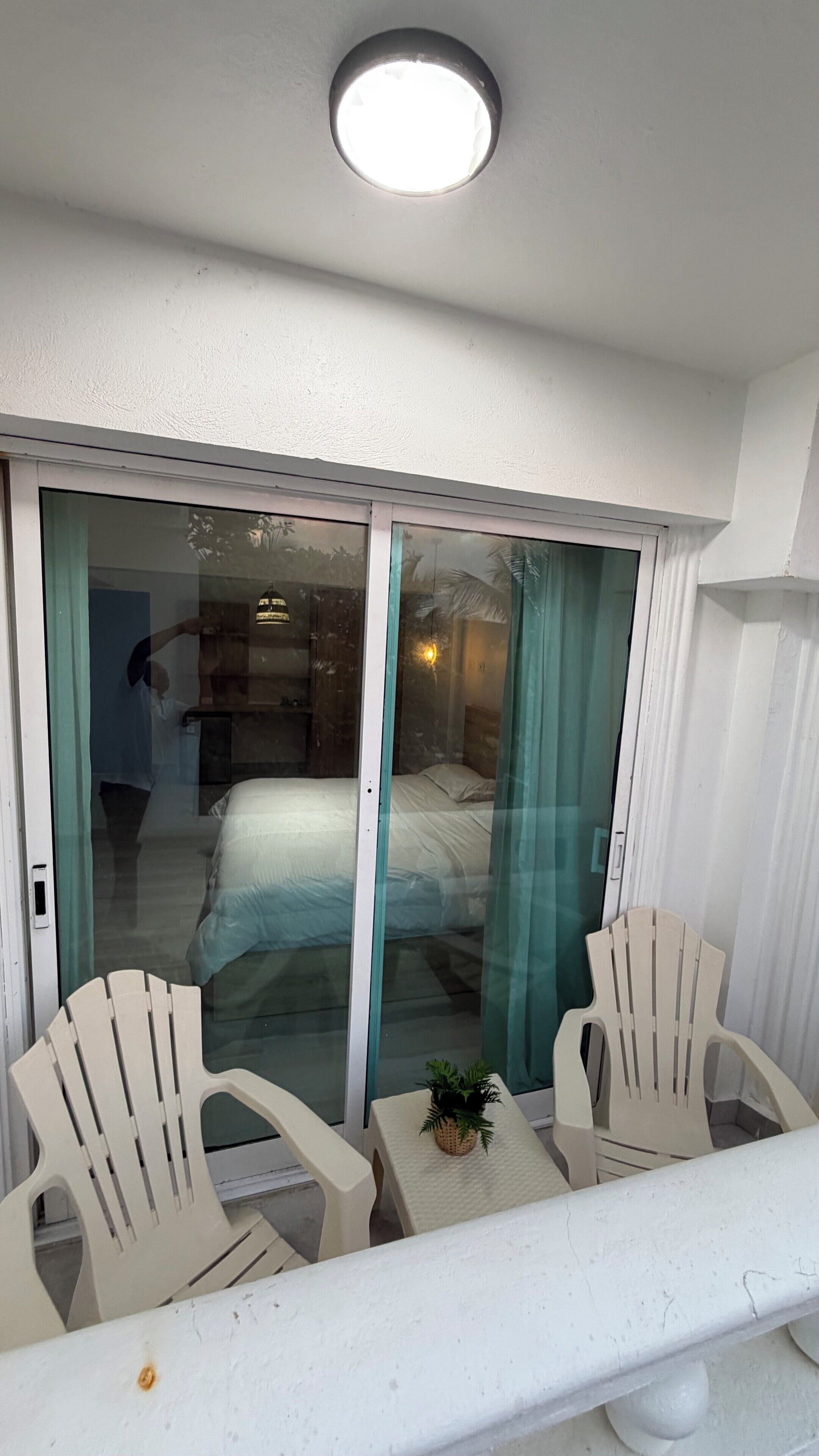 Varanda com cadeiras brancas, uma mesa pequena e uma porta de vidro deslizante que dá para um quarto com cama e mesa de cabeceira.