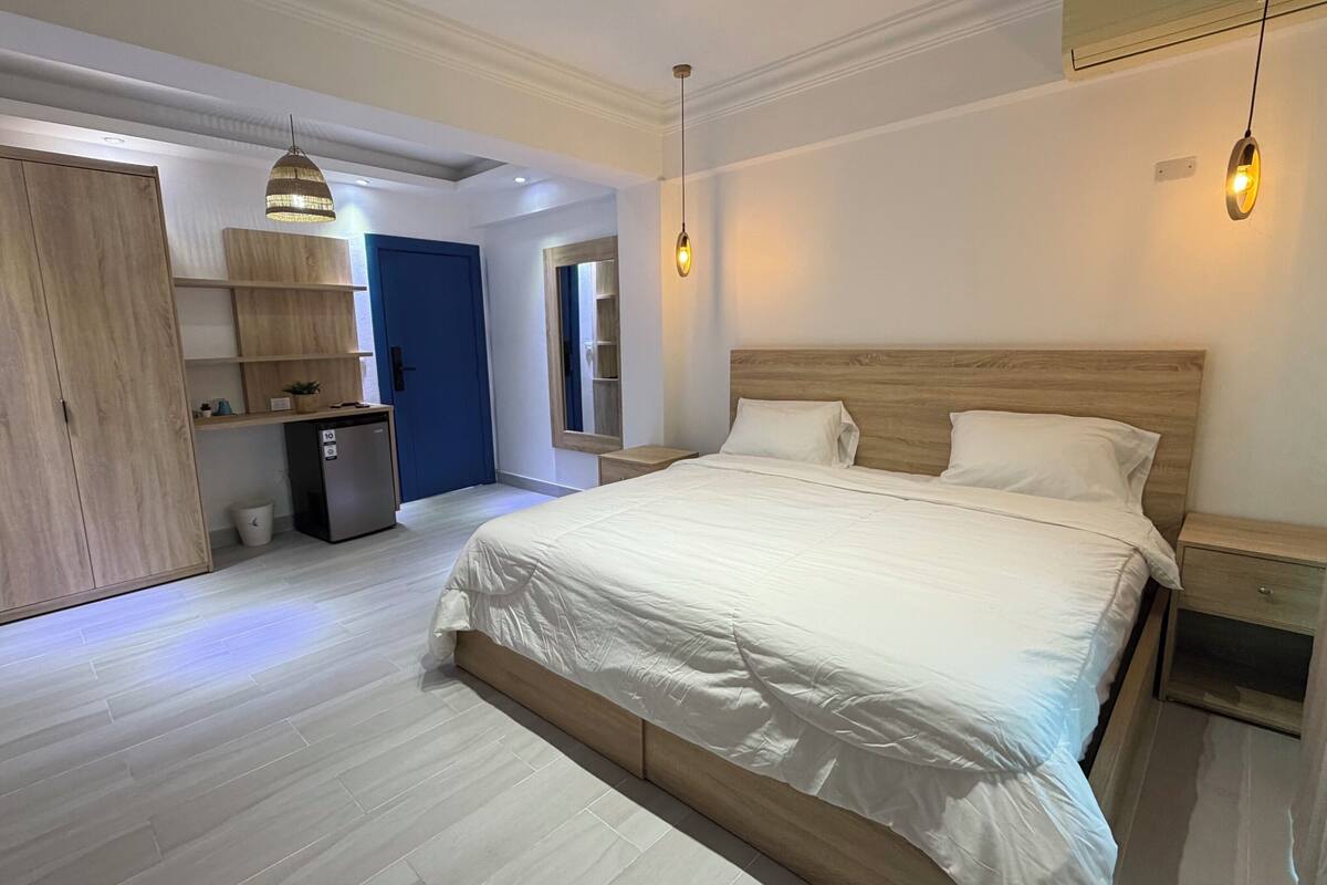 A modern bedroom with a large bed, wooden headboard, and bedside tables. There is a small kitchenette with a refrigerator and a sink. A blue door leads to another room.