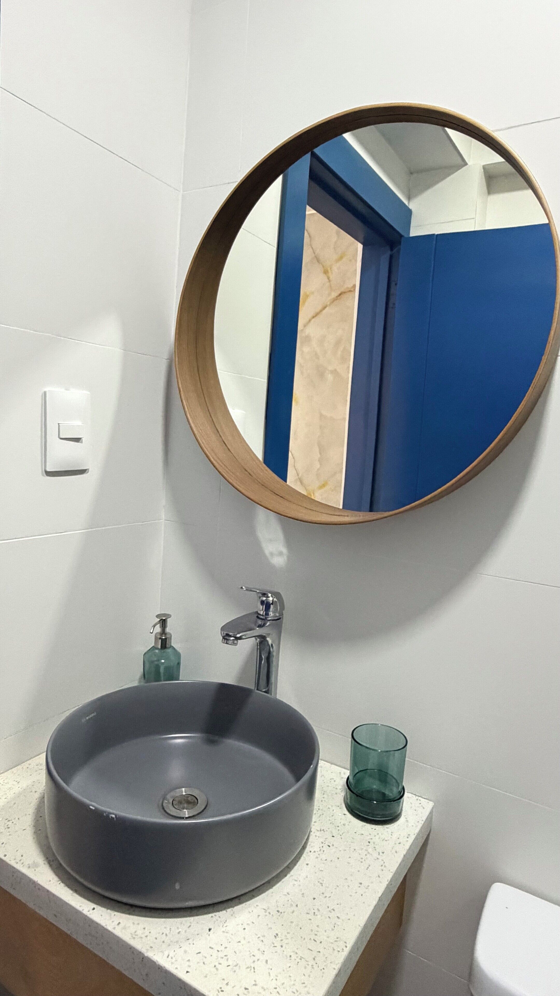 Un baño con un espejo redondo, un lavamanos, un grifo, dispensador de jabón y un vaso.