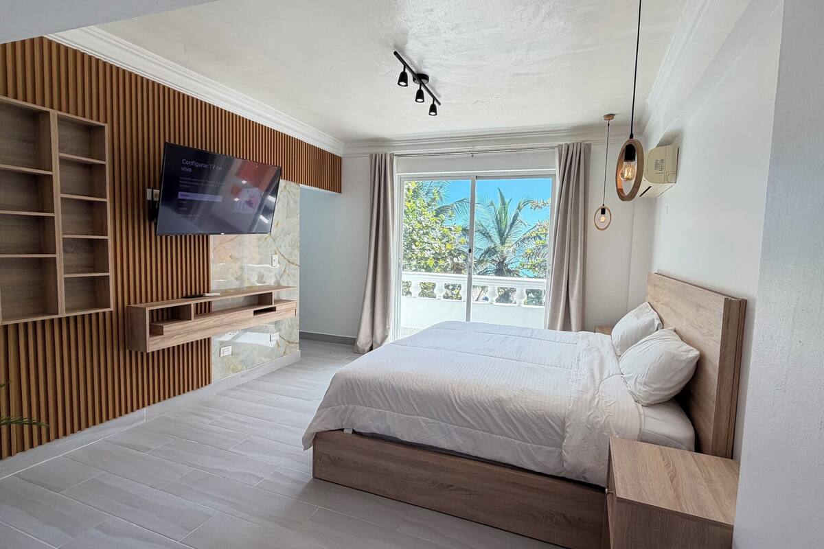 A bedroom with a large bed, a TV mounted on a wooden panel wall, a balcony with a view of trees, and a ceiling light fixture.