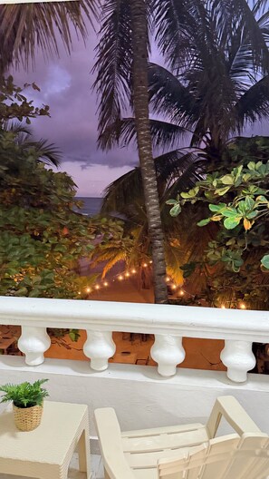 A balcony with a white balustrade, a wicker table with a potted plant, and a view of palm trees and string lights against a twilight sky. - Hotel Juan Dolio (Guayacanes)
