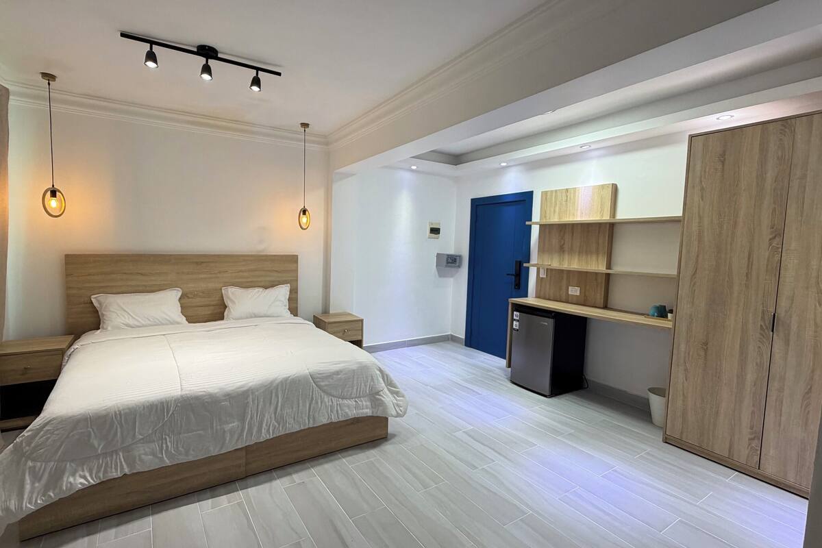 A modern hotel room with a large bed, bedside tables, a wardrobe, and a small kitchenette.
