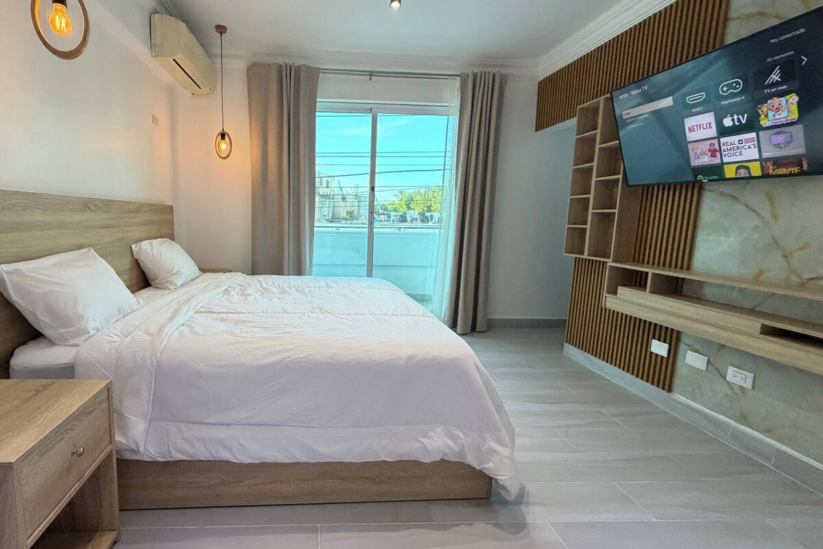 A modern hotel room with a large bed, wooden headboard, a TV mounted on the wall, and a view of the outside through the window.