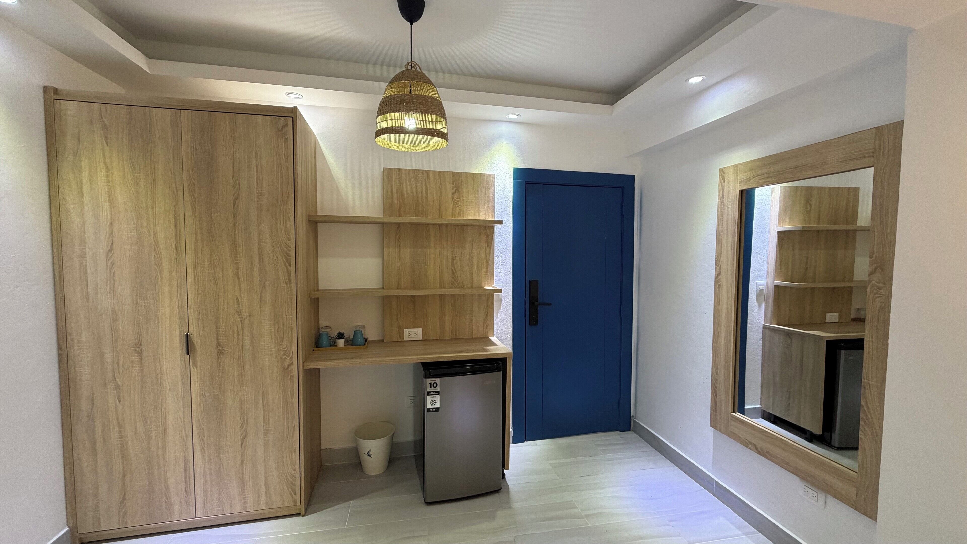 A hallway with a wooden wardrobe, a small refrigerator, and a blue door. Shelves are mounted on the wall, and a hanging light fixture is visible.