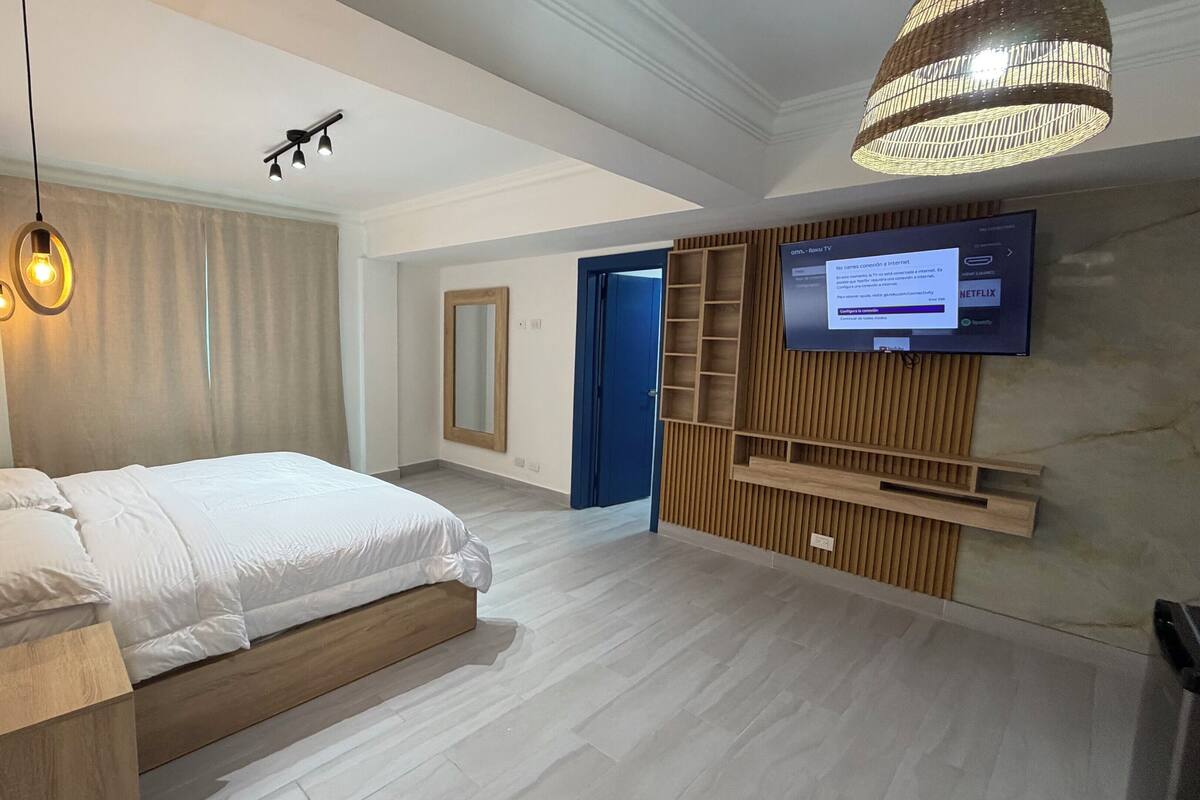 A modern bedroom with a bed, wooden headboard, a TV mounted on the wall, and a ceiling light fixture.
