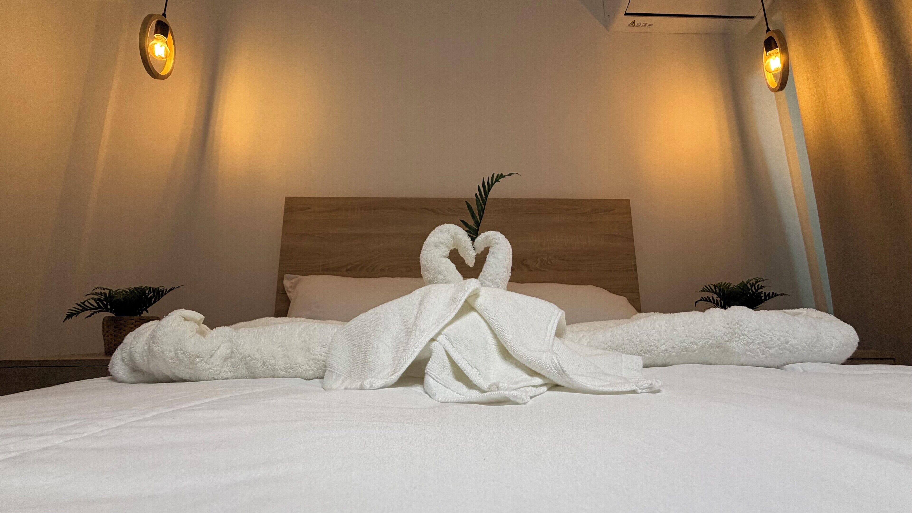 A neatly made bed with a heart-shaped towel arrangement, a wooden headboard, and two bedside plants.